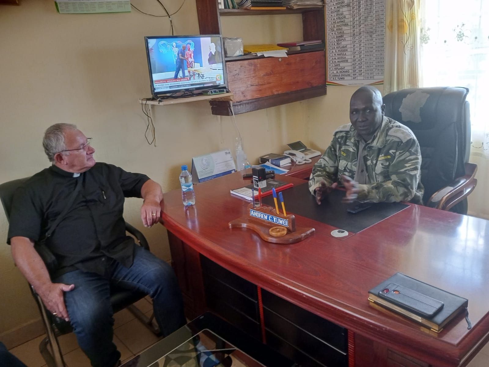 Two men are sitting at a desk in front of a television.