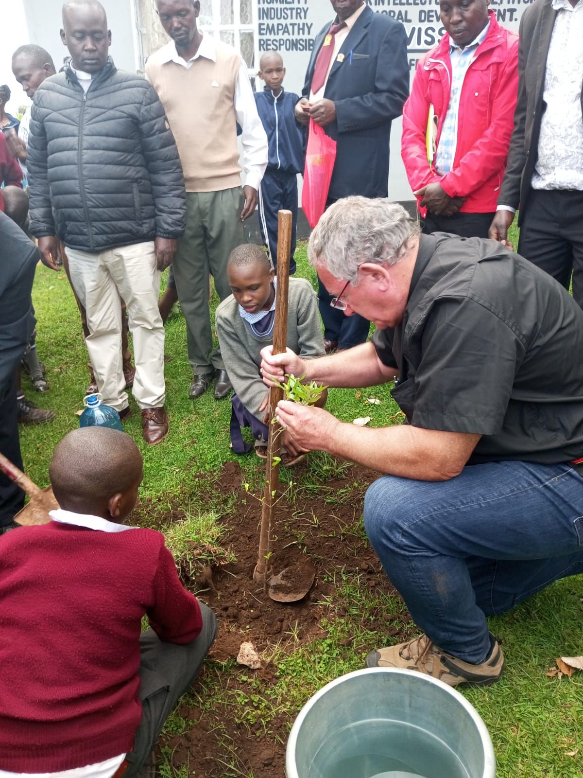 A man is kneeling down to plant a tree while a group of people watch.