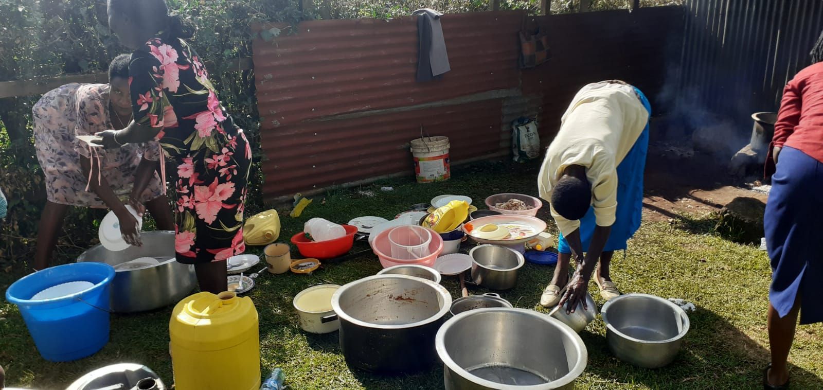 A group of people are standing around pots and pans on the grass.