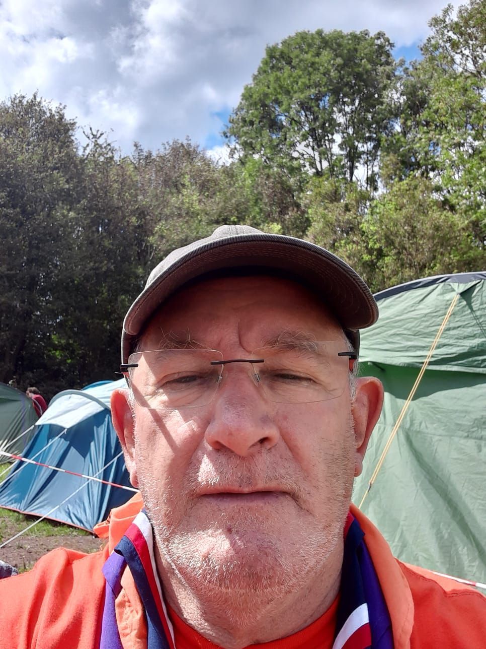 A man wearing glasses and a hat is taking a selfie in front of tents.