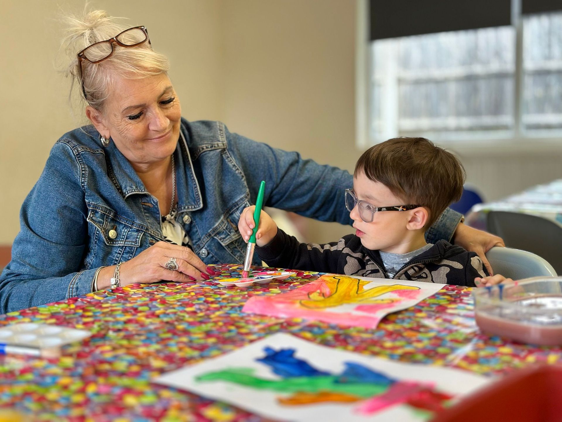A woman is helping a young boy paint at a table.