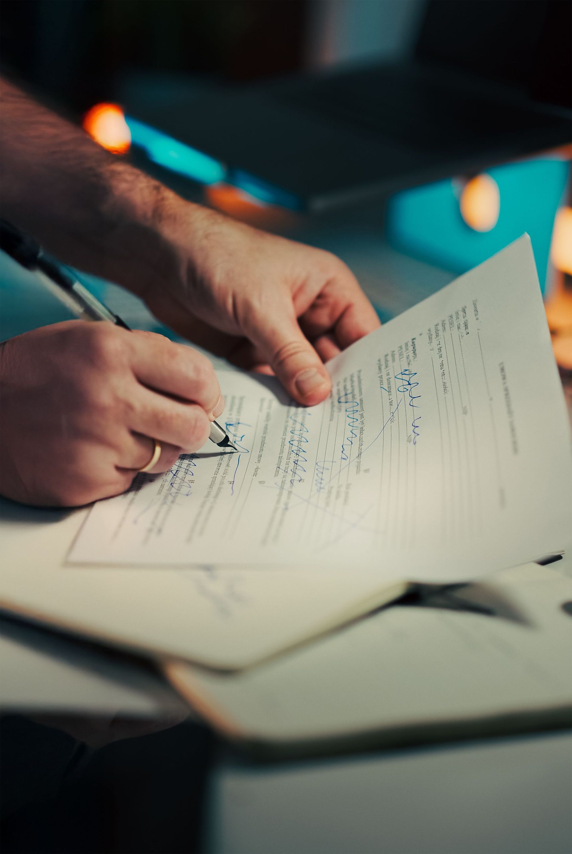 Hands writing on a printed form at a desk, with blurred lights in the background.