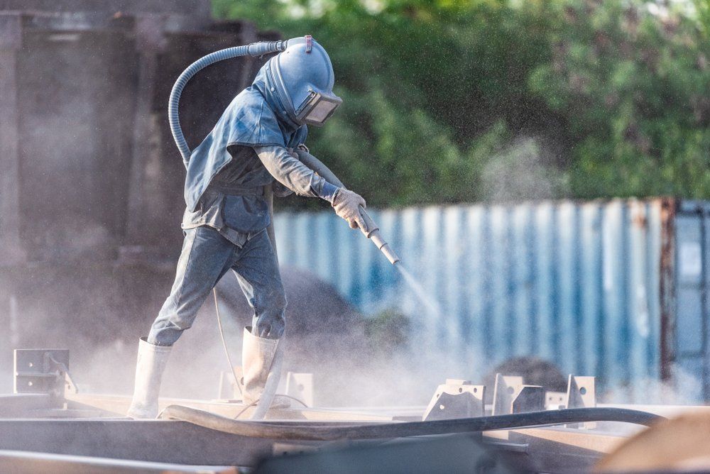 A Man in A Protective Suit Is Sandblasting a Piece of Metal — Mayric Engineering in Hallidays Point, NSW