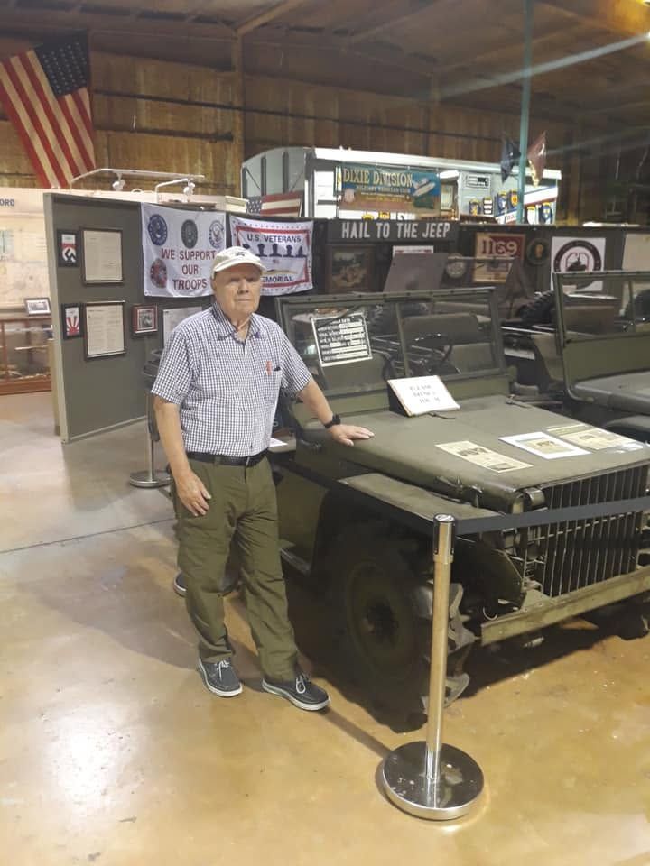 Man standing beside a military jeep in a museum display, with informational signs and an American flag in the background
