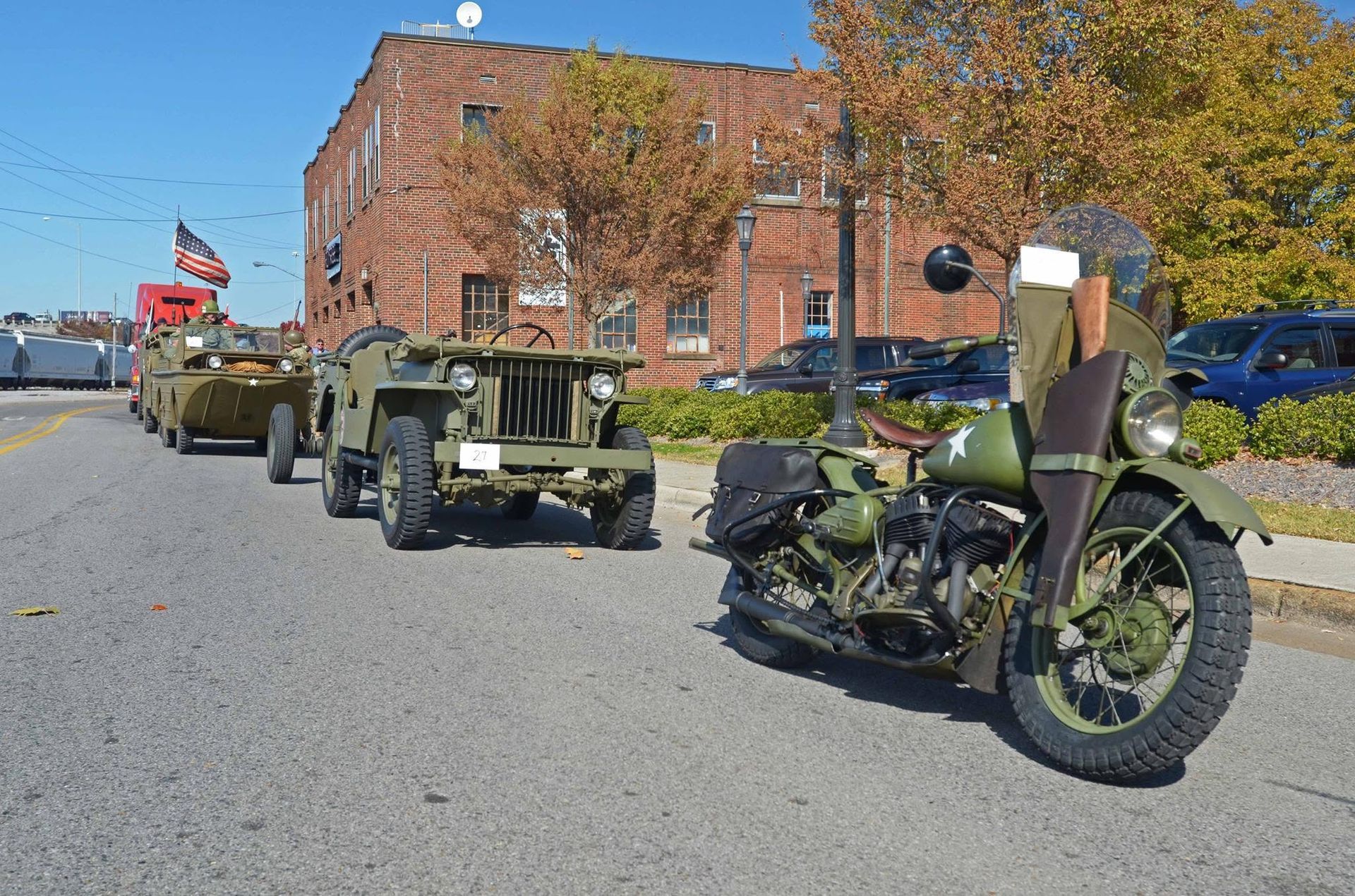 Military jeep towing a green motorcycle on a street beside a brick building and parked cars