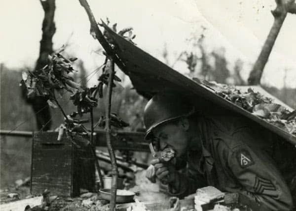 Soldier crouched in a small trench beneath camouflage netting, surrounded by bare trees and muddy ground