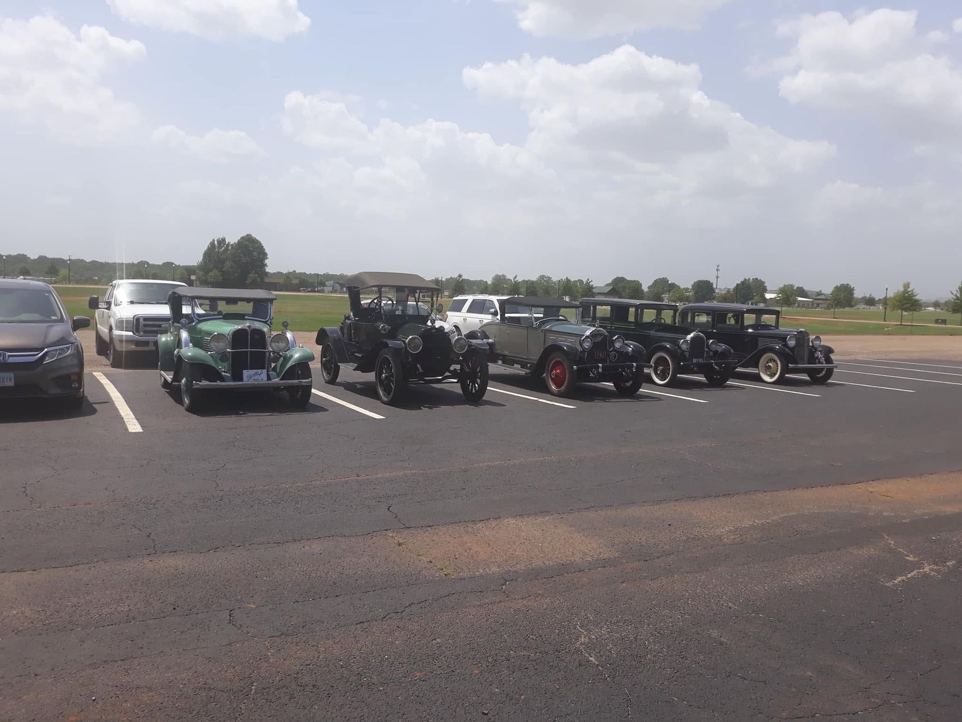 Several vintage cars parked in a lot under a cloudy sky, with people standing by them