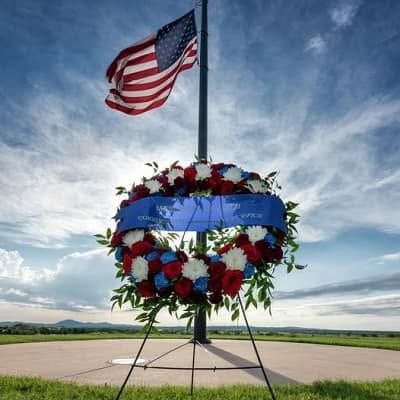 American flag above a memorial wreath with red, white, and blue flowers against a cloudy sky