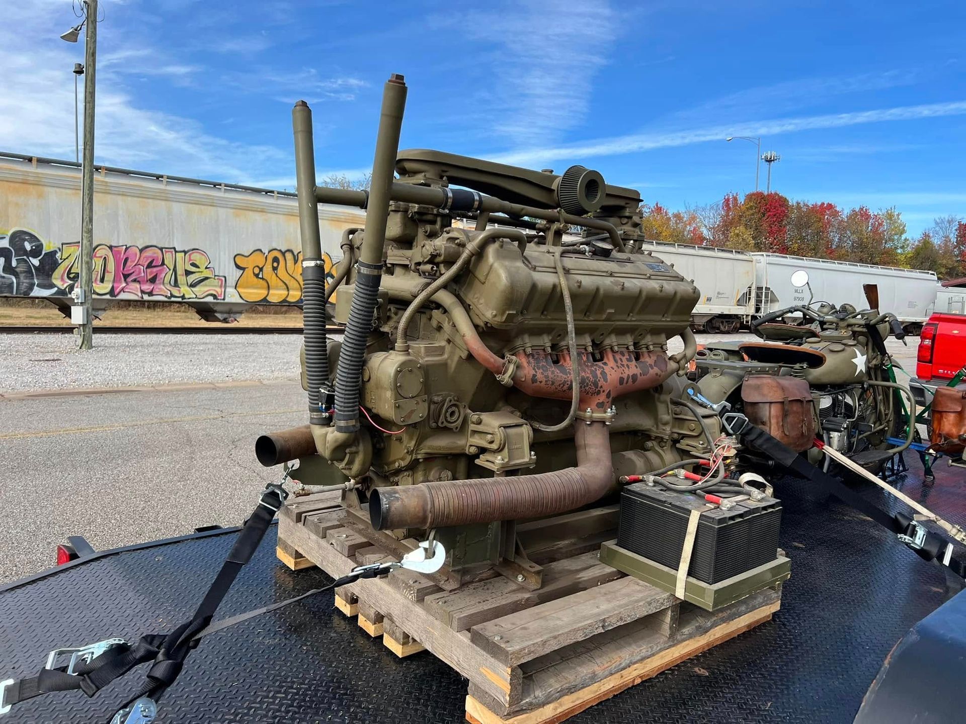 Large old engine strapped to a trailer in a parking lot, with graffiti wall and autumn trees behind it