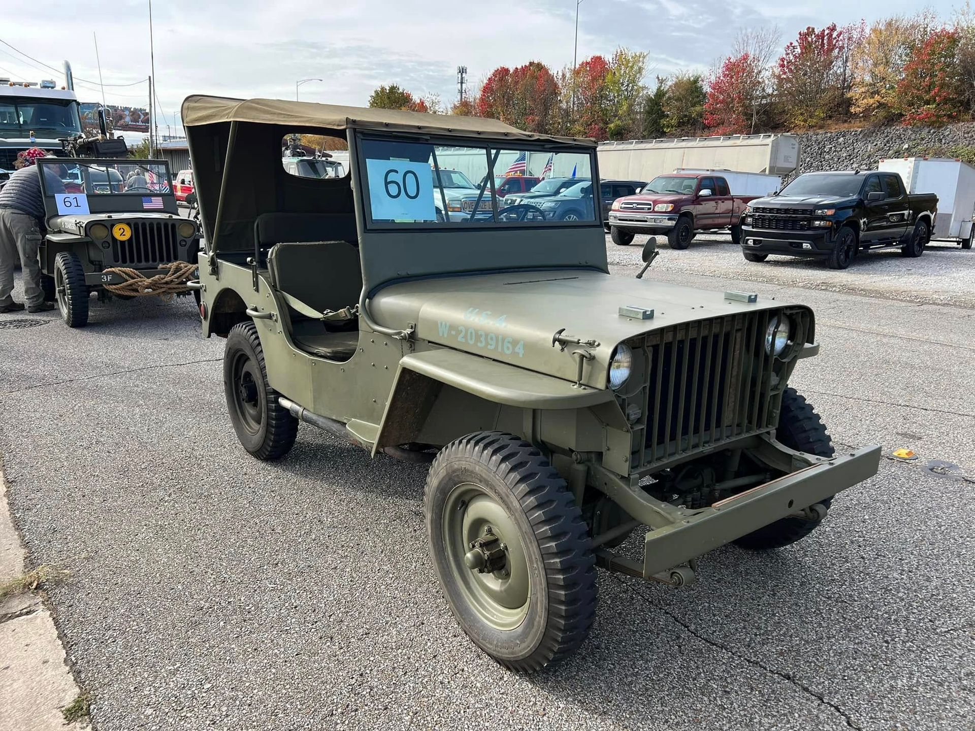 Olive drab vintage military jeep parked on gravel at an outdoor car show, with a blue number card on the windshield