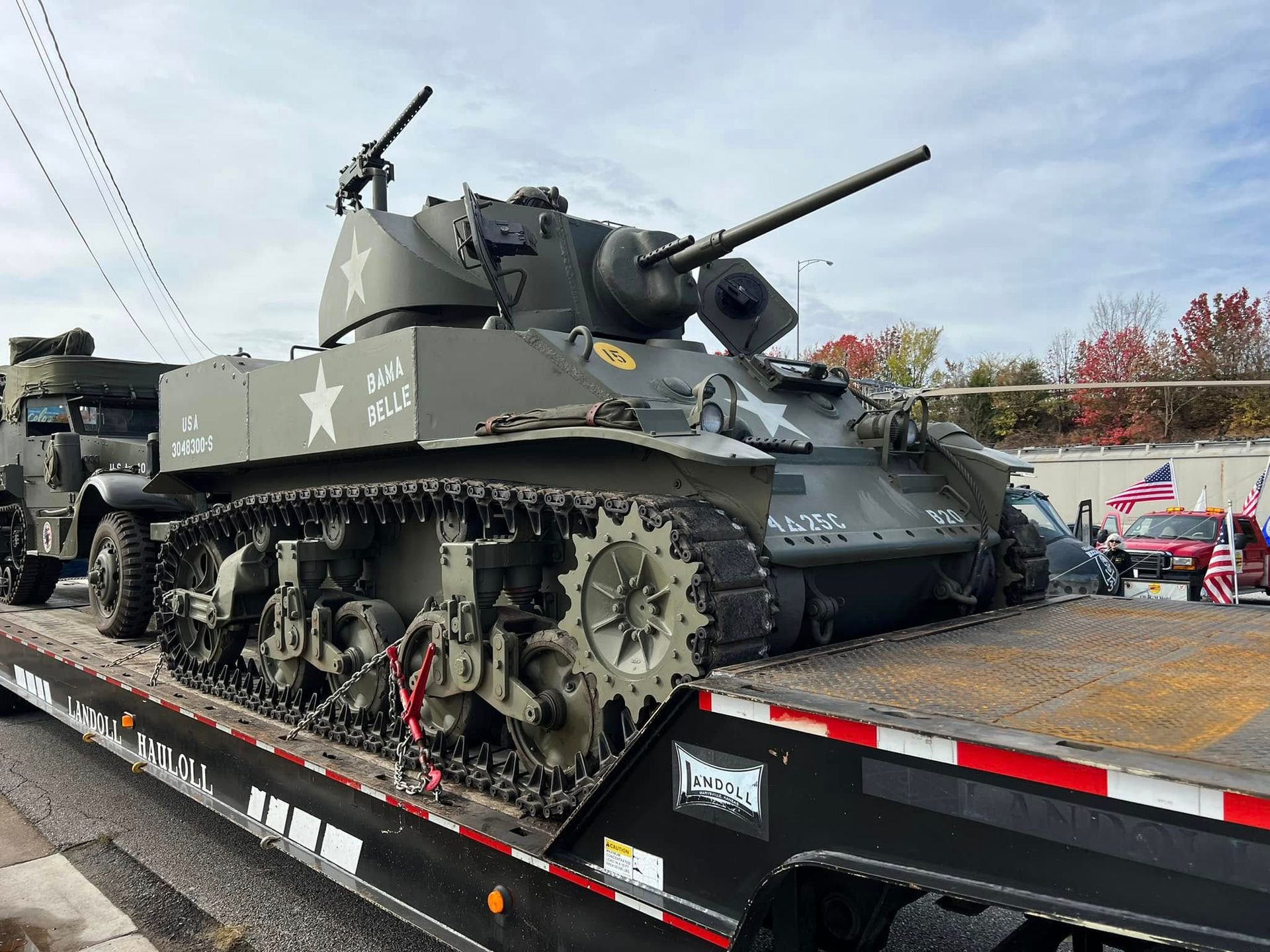 Gray tank on a trailer at an outdoor display, with a military vehicle in the background and autumn trees.