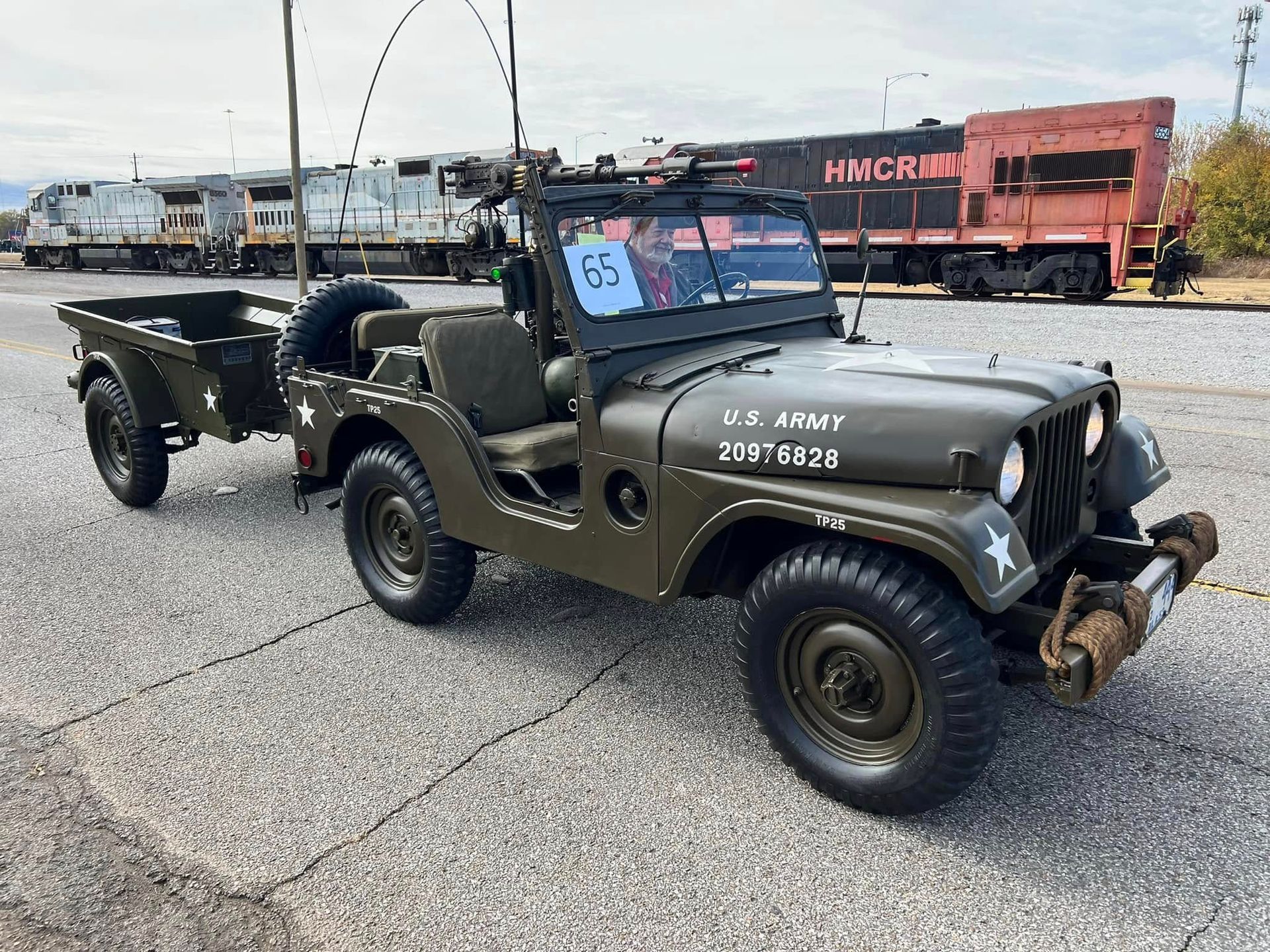 Military-style olive jeep with trailer parked on a gravel lot, front view.