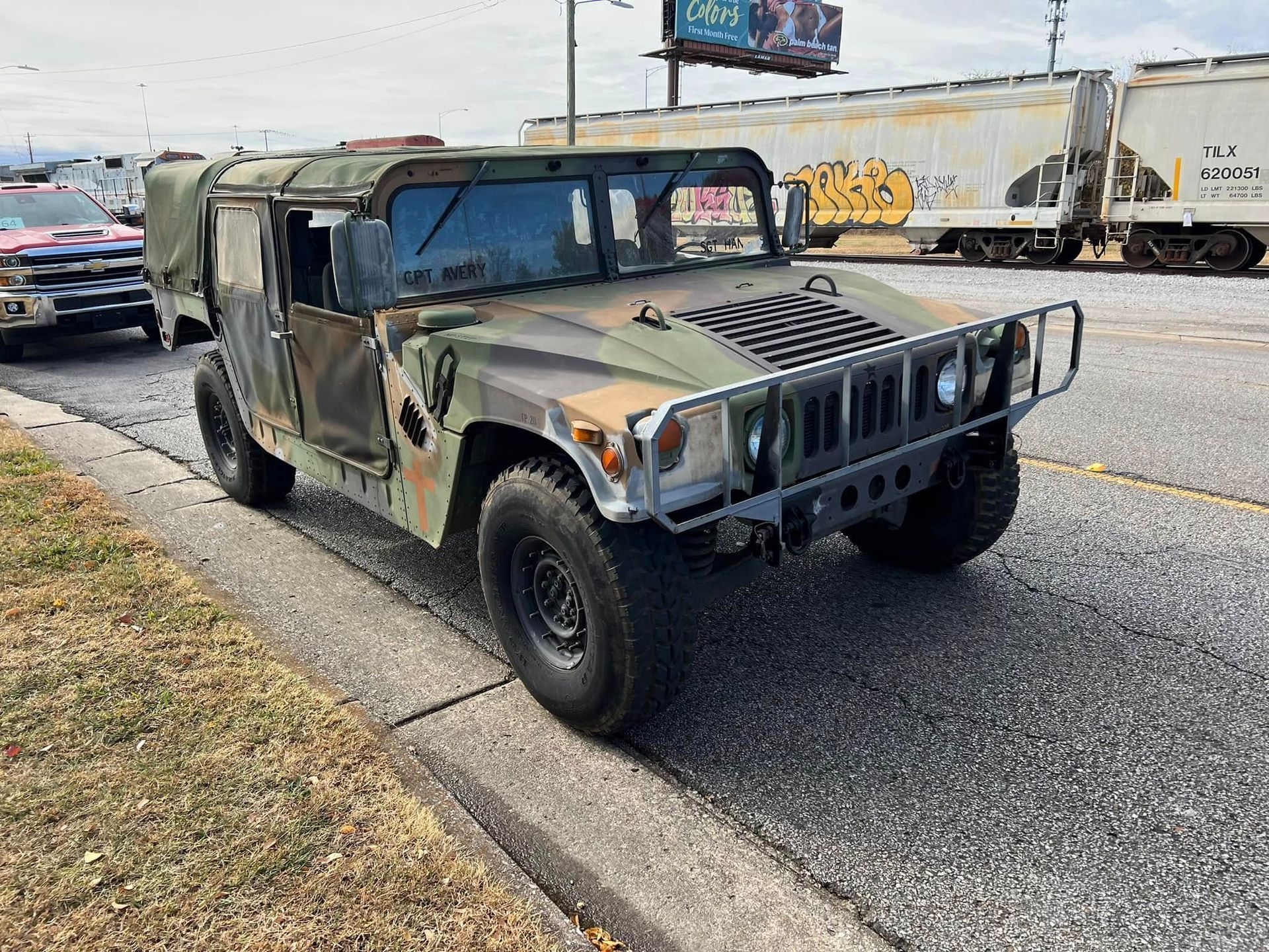 Camouflaged military Humvee parked on a roadside beside train tracks and a billboard.