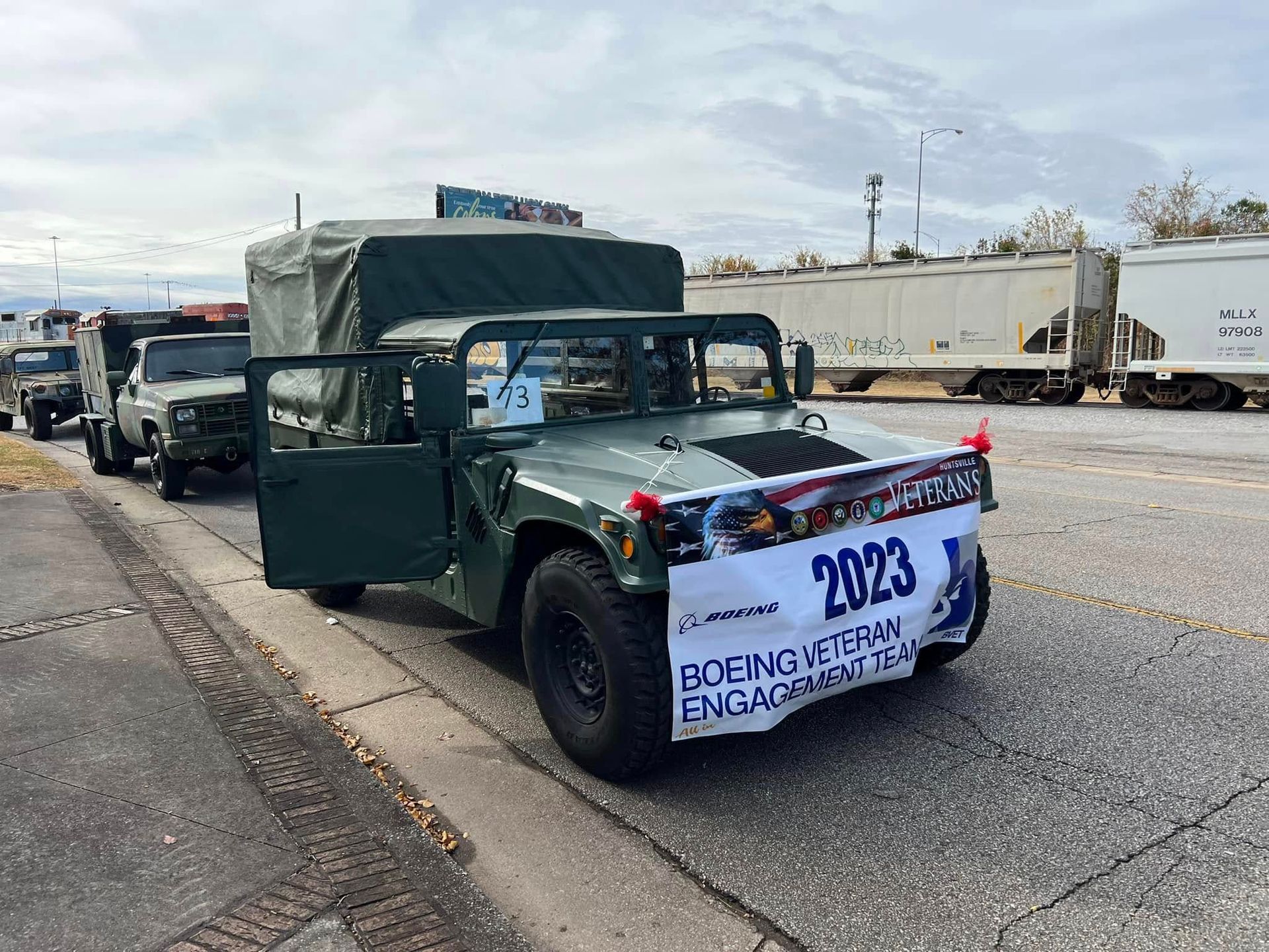 Green vintage truck decorated with a “2023” banner, parked on a street near other vehicles and trailers.