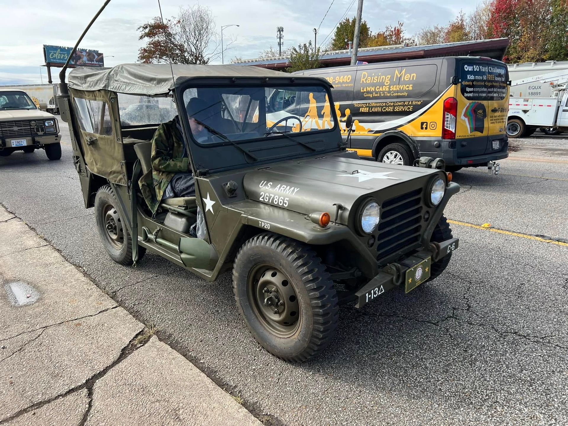 Olive drab military jeep parked on a street beside a black-and-yellow van with graphics