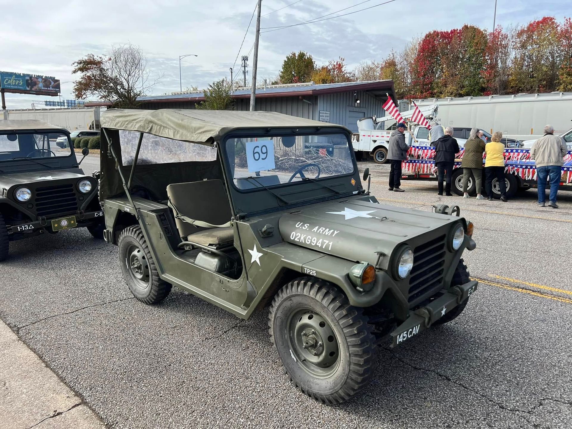 Olive drab vintage military jeep with canvas top parked on gravel at a roadside event, people in background
