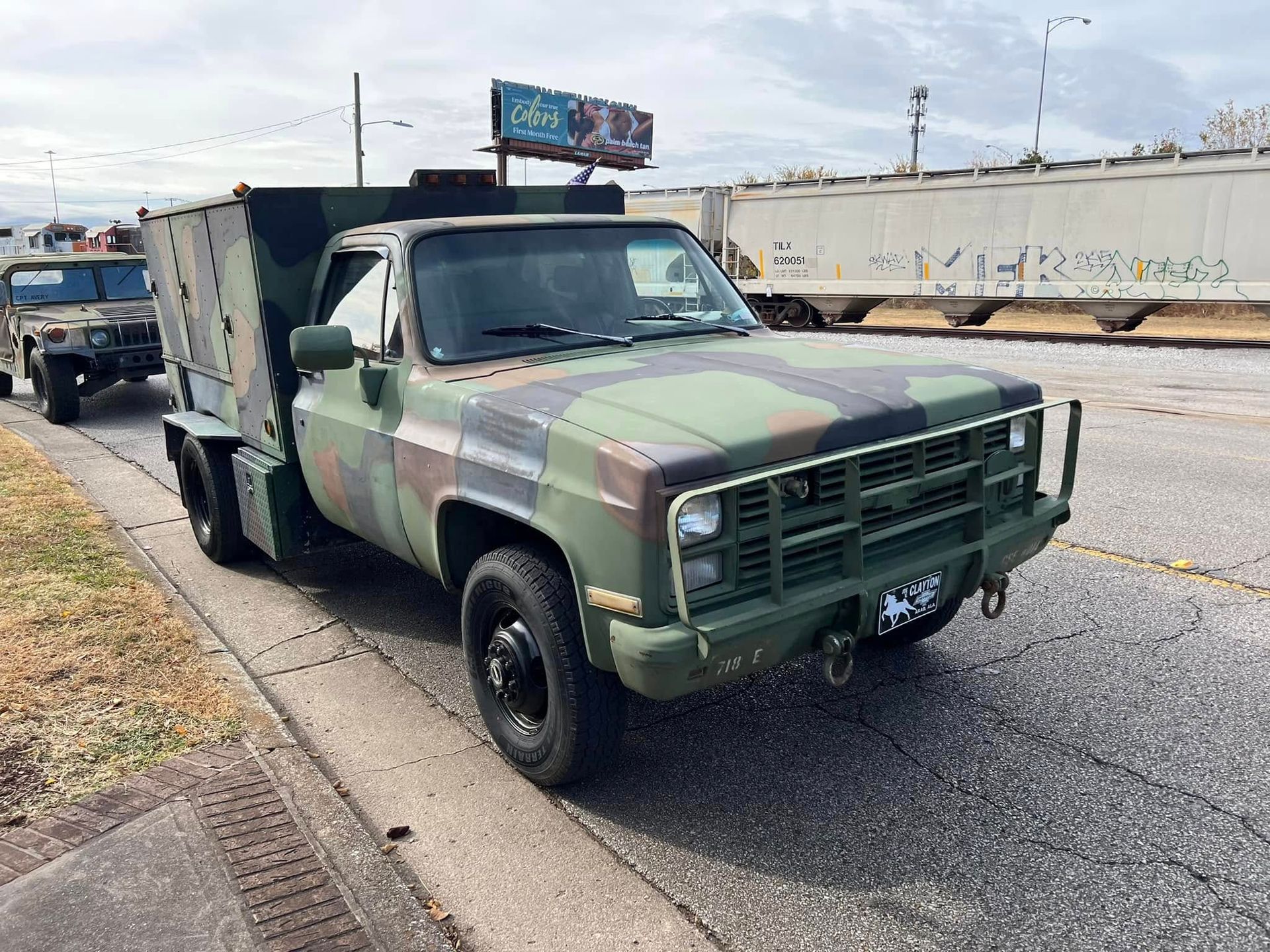 Camouflage pickup truck parked on a roadside with a trailer attached near railroad tracks.