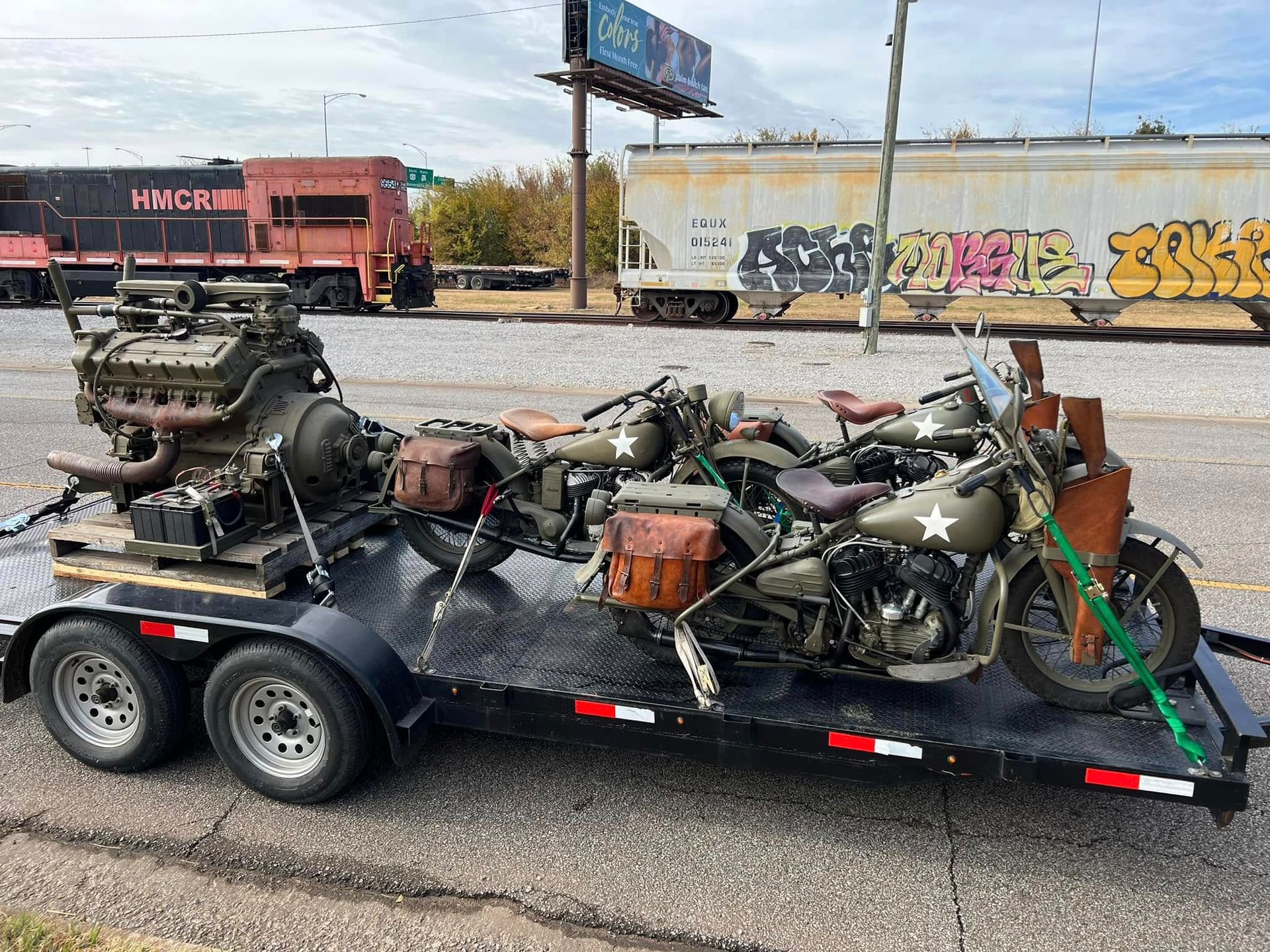 Black dual-axle trailer loaded with old vehicle engines and parts beside a graffiti-covered wall.