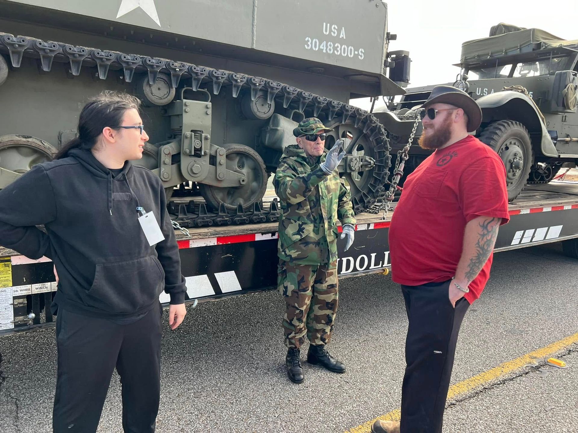 People standing beside a military truck on a road, with an armored vehicle and camouflage-covered cargo in the background.