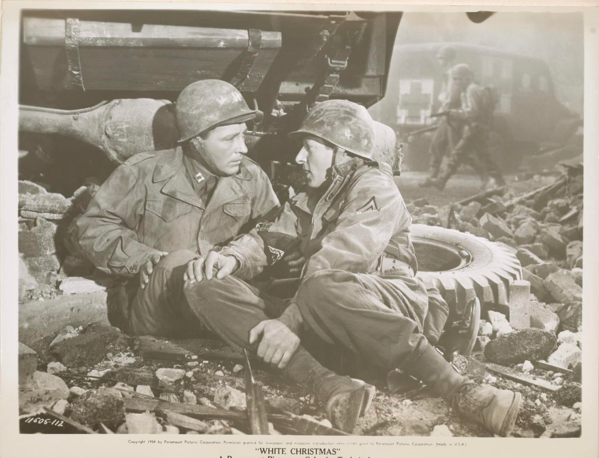 Two soldiers in helmets sit amid rubble, talking beside a damaged vehicle in a war zone.