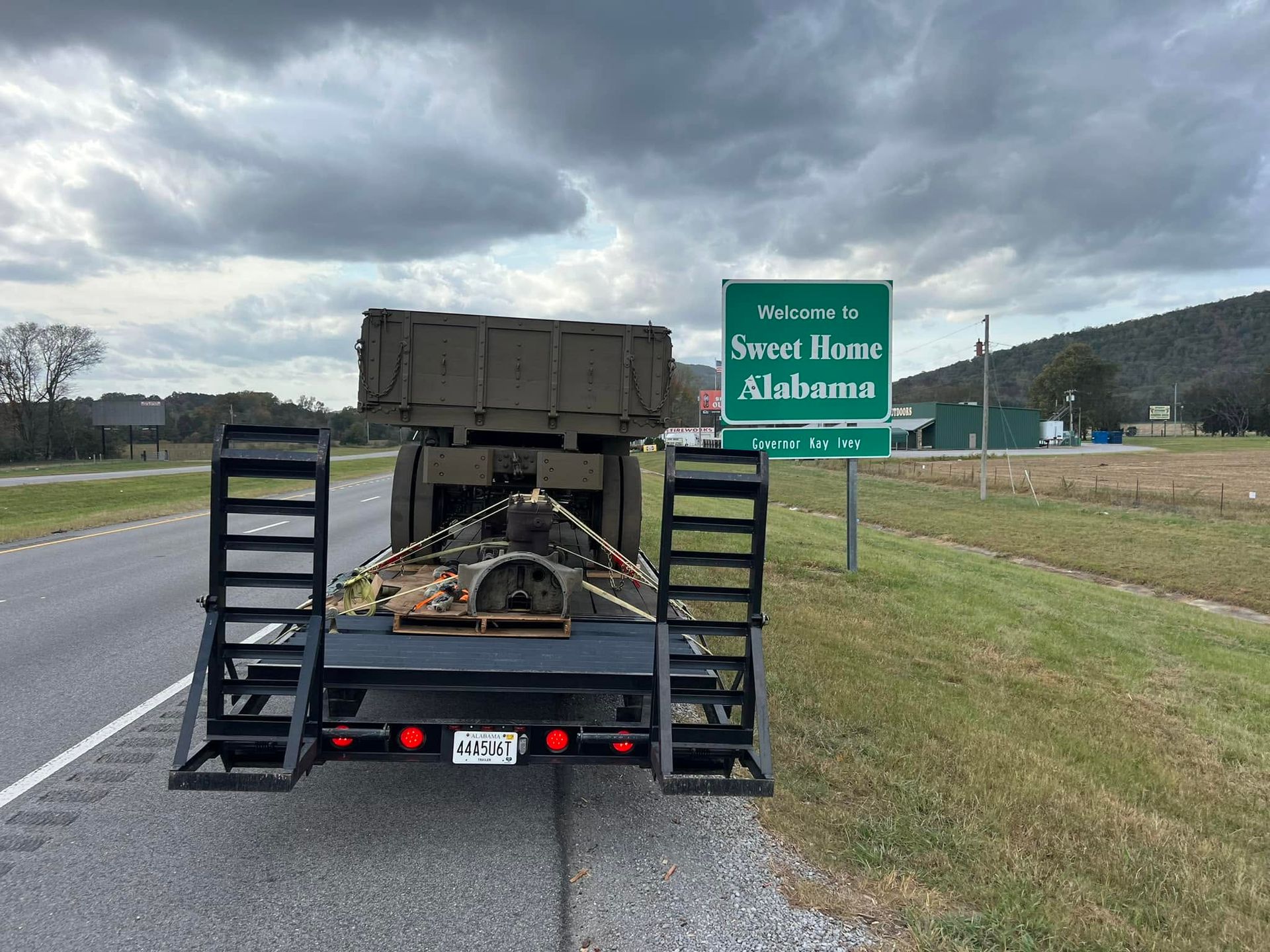 Trailer hauling debris on roadside near a green “Welcome to Sweet Home Alabama” sign under cloudy skies