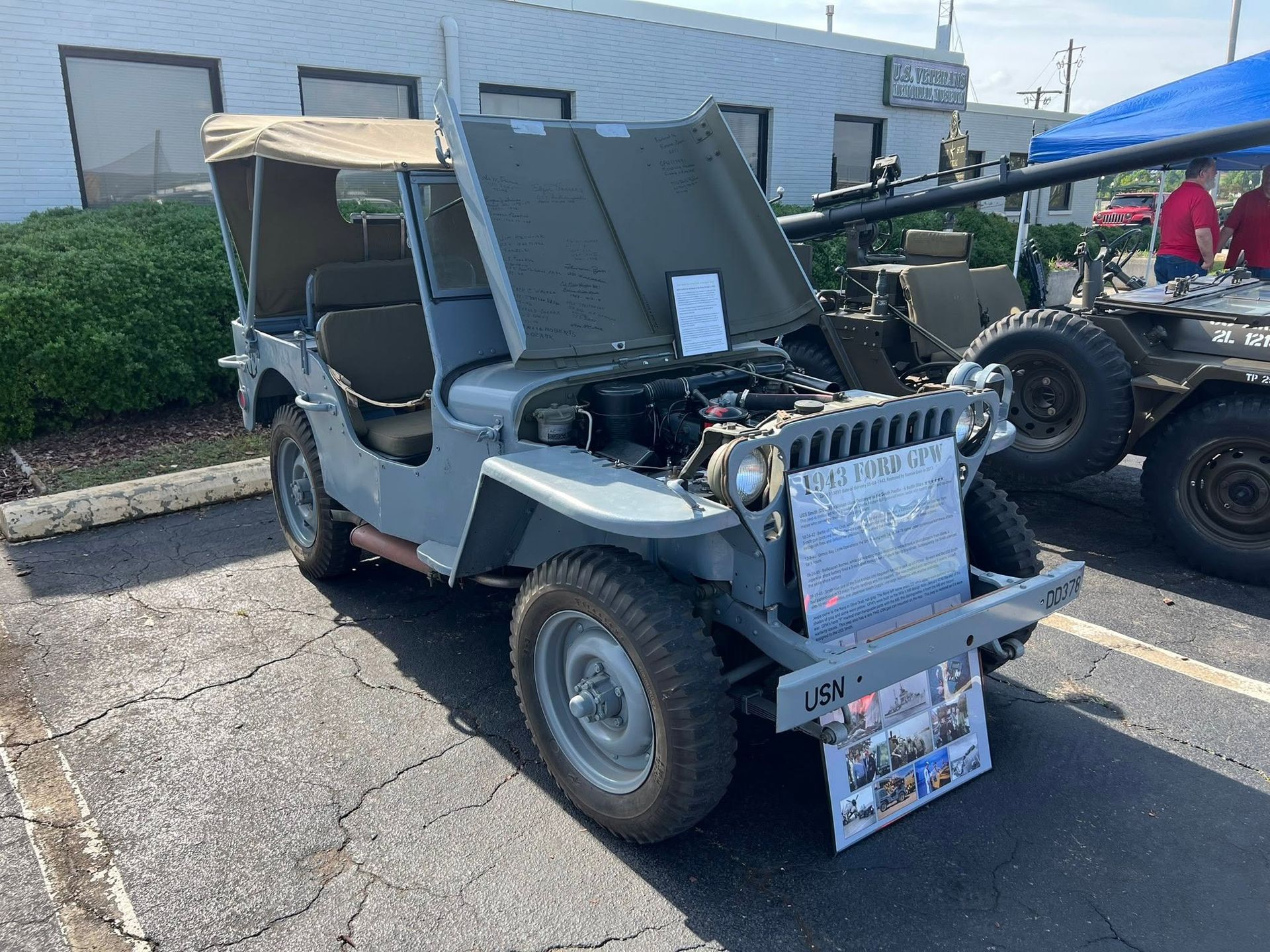 Vintage gray off-road jeep on pavement at an outdoor display, with open hood and sign on the front.