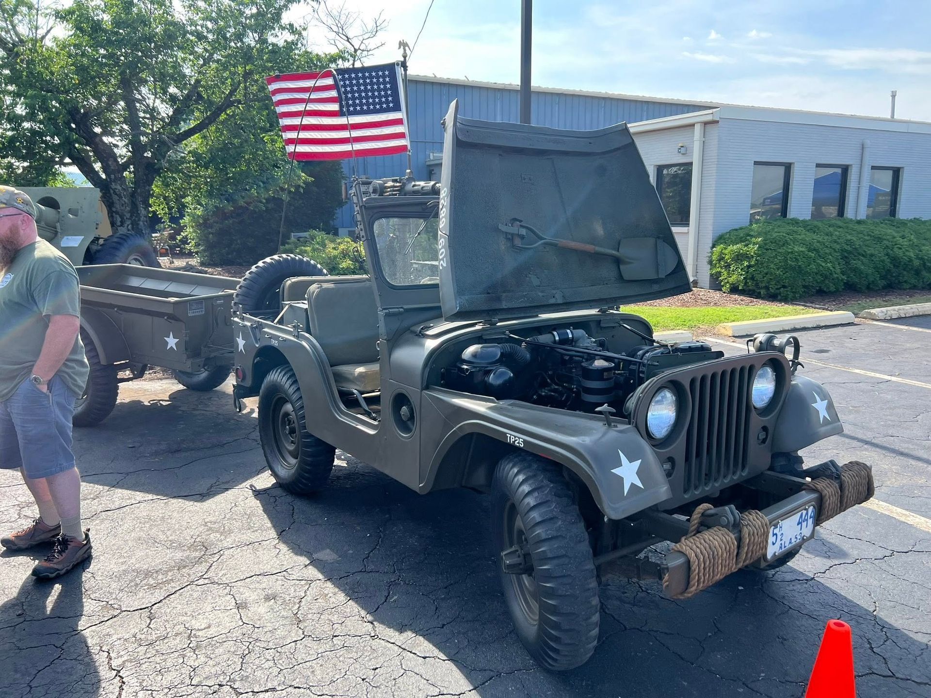 Military jeep with open hood and American flag parked in a lot beside a man and trailer