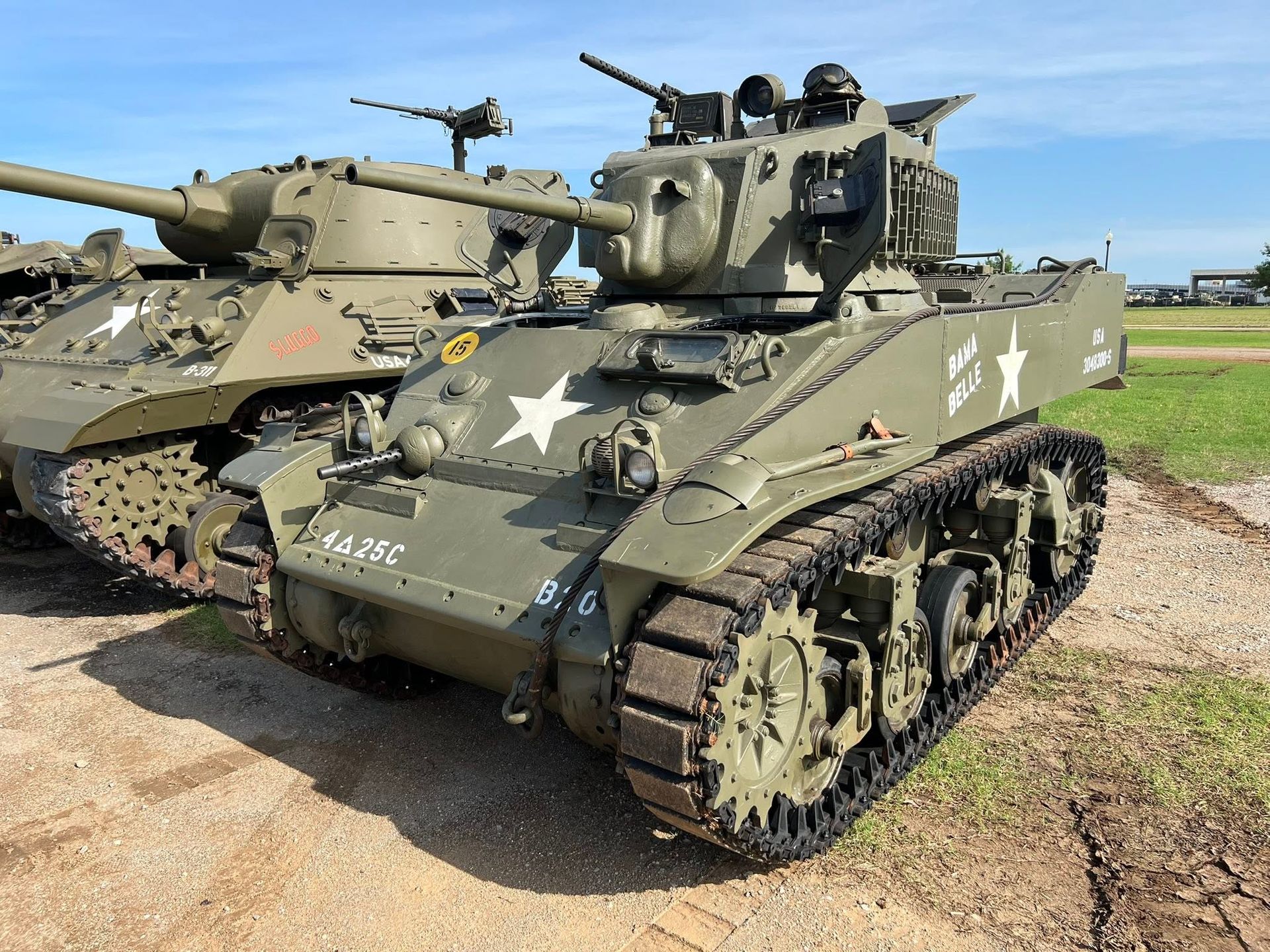Two camouflaged military tanks with mounted guns parked on grass under a blue sky.
