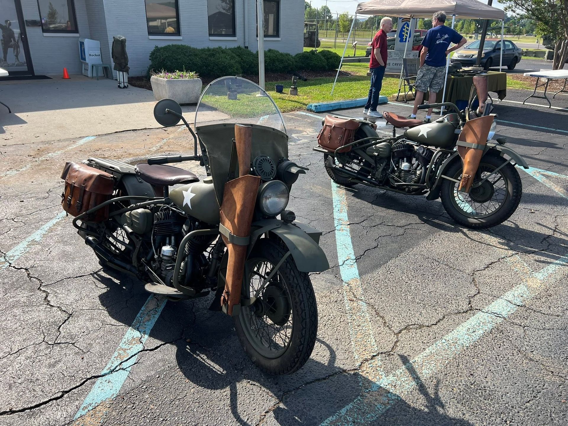 Two vintage motorcycles parked in a lot, one with a sidecar, near a building and people in the background