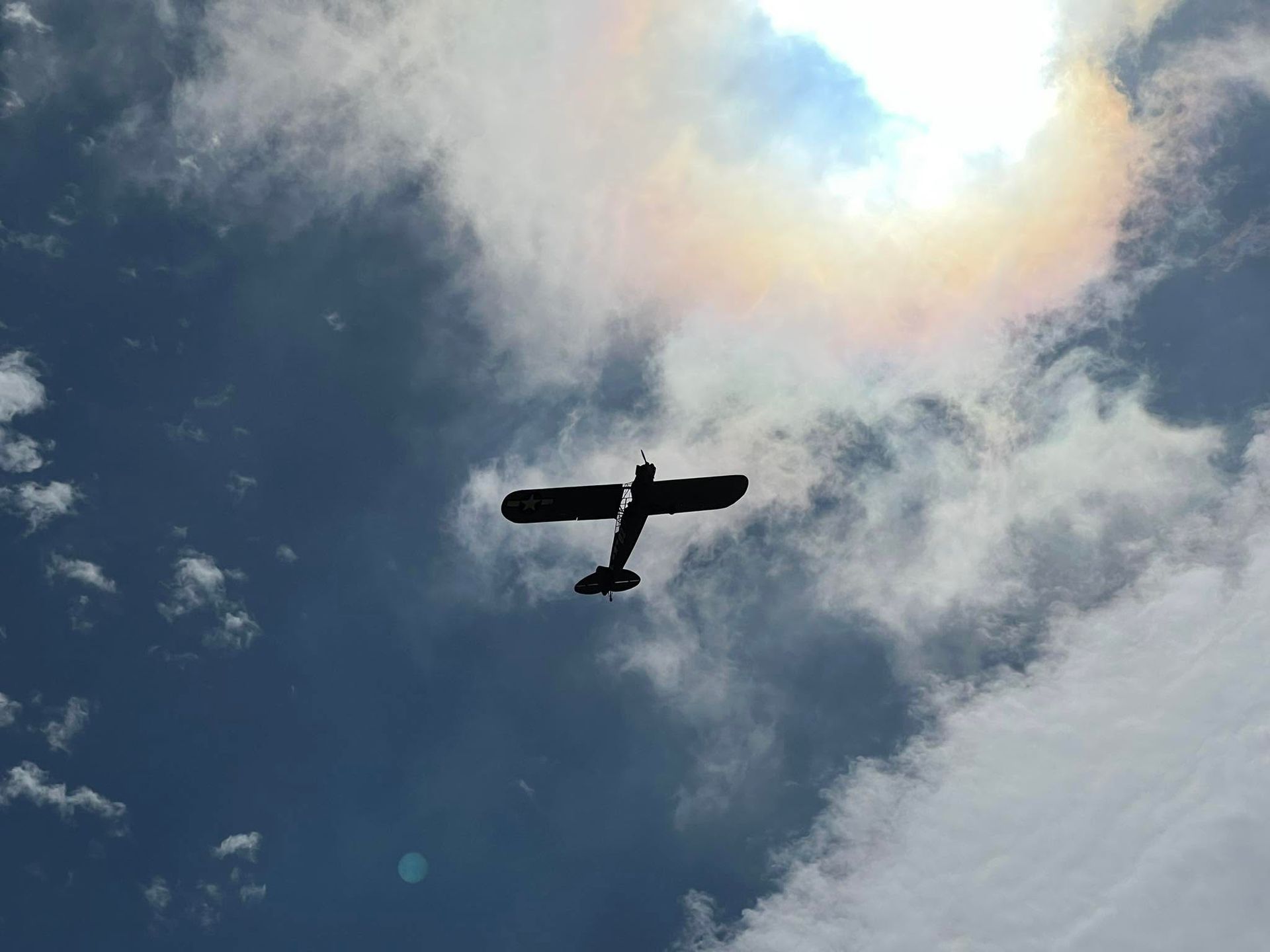 Silhouette of a small airplane flying against a cloudy blue sky with a bright sun halo