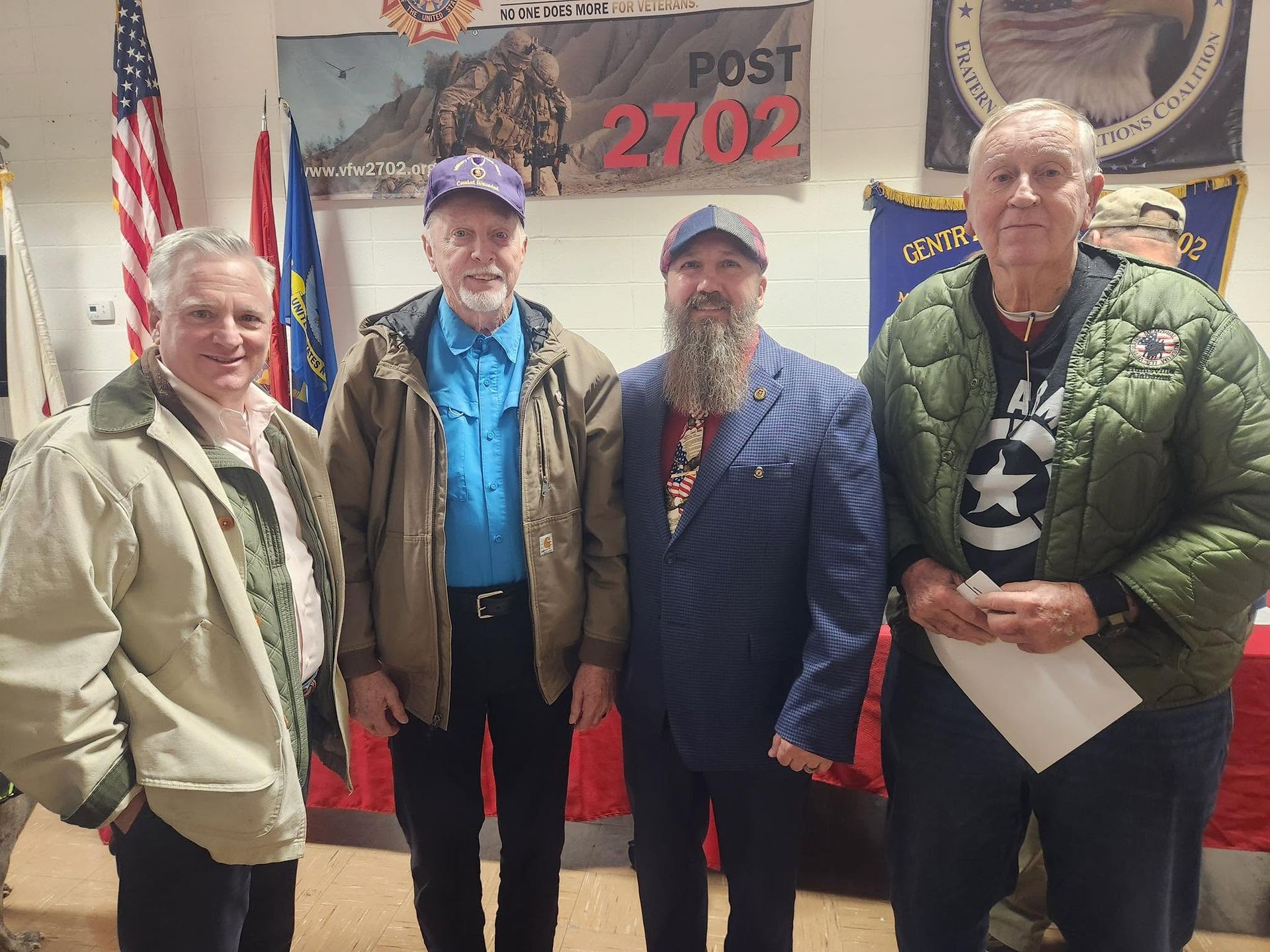 Four men pose indoors in front of a VFW Post 2702 banner and flags.