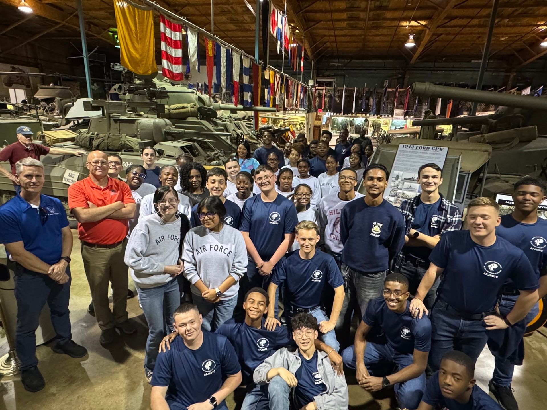 Group of people posing in a hangar beside a military aircraft, with flags hanging overhead.