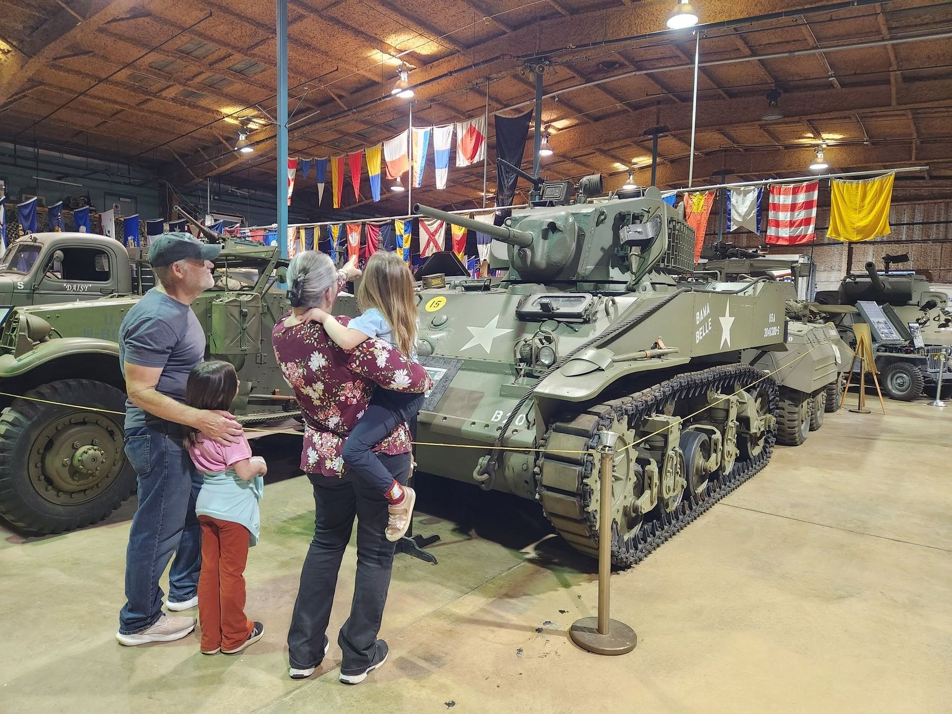 People viewing a gray military tank in a museum, with flags hanging overhead.