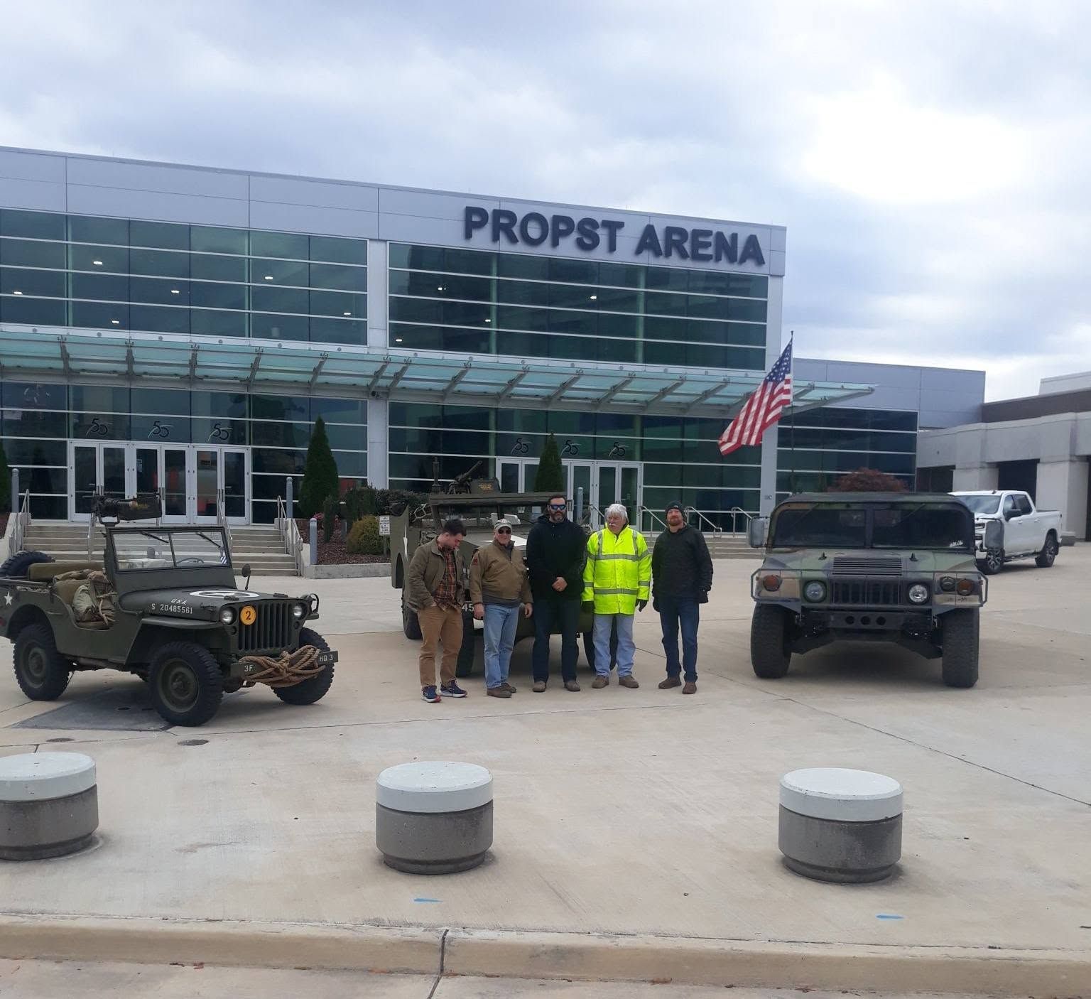 People in reflective vests stand outside Propst Arena beside military vehicles and an American flag.
