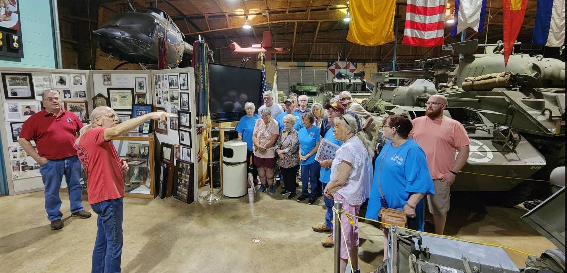 Group of people posing in a museum beside military vehicles and flags, some waving at the camera.