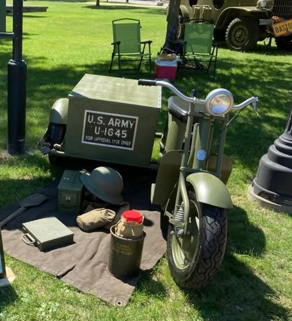 Olive drab vintage military scooter with U.S. Army sign displayed on grass outdoors.
