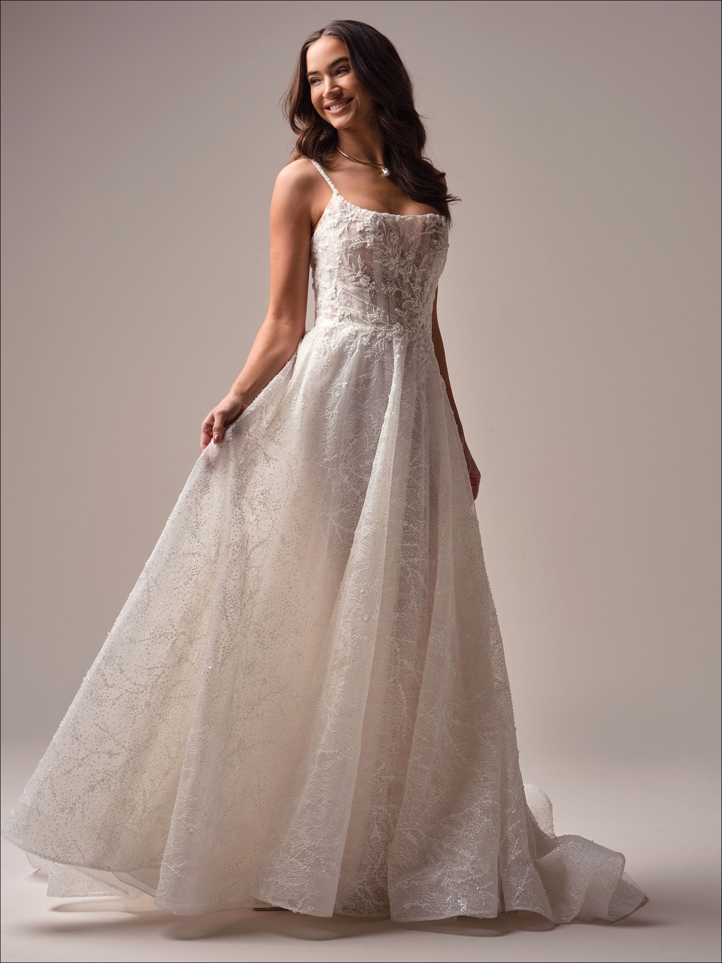 Woman in a wedding dress, smiling, holding out the skirt, in a studio setting.