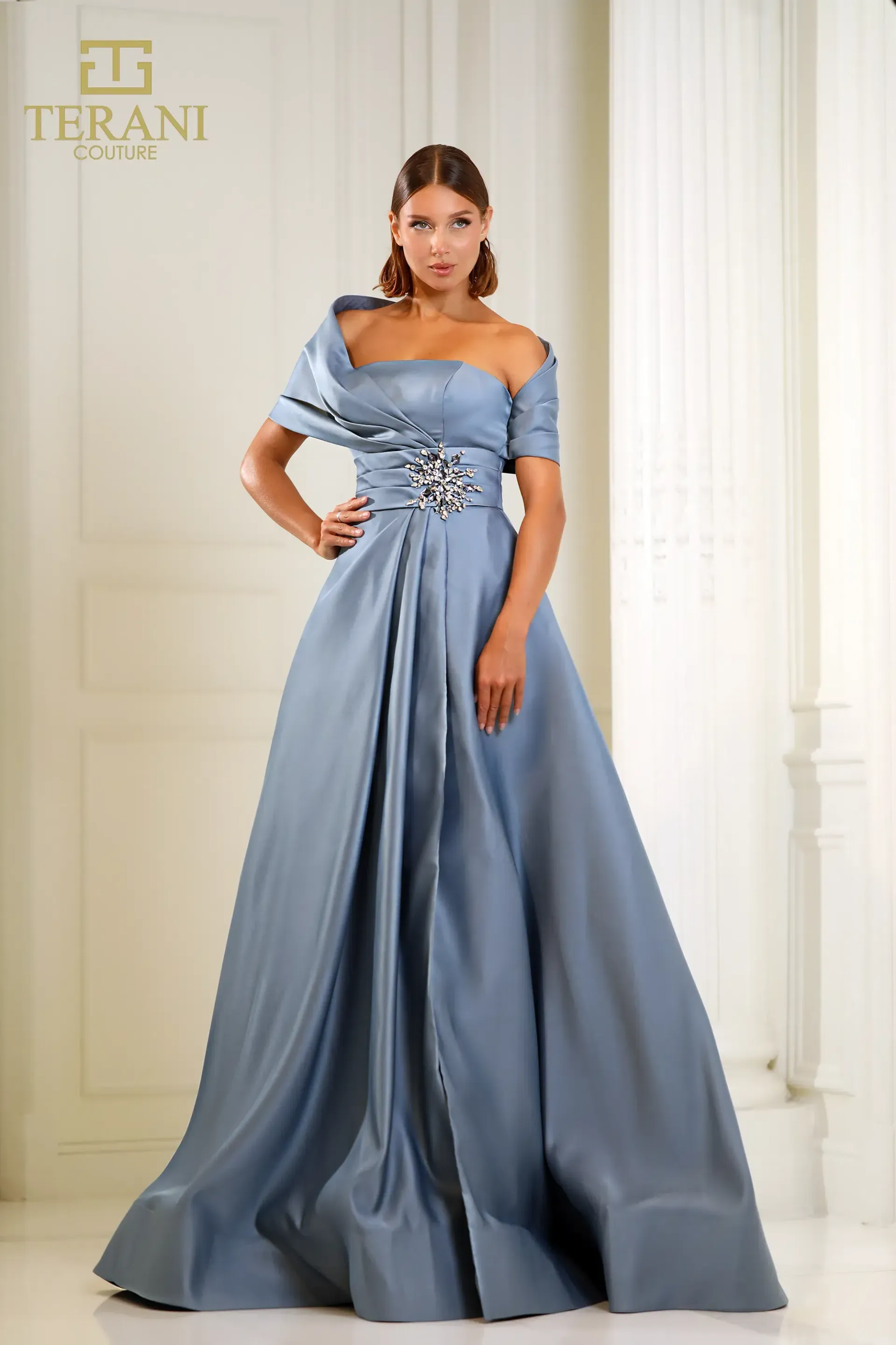 A woman in a long blue dress is standing in front of a white wall.