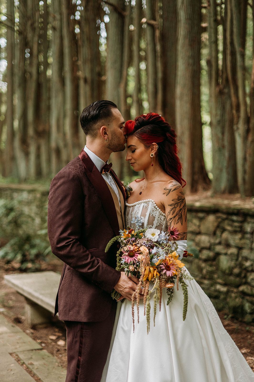 Groom kisses bride's forehead in forest. Bride in white dress, holding bouquet. Groom wears burgundy suit.