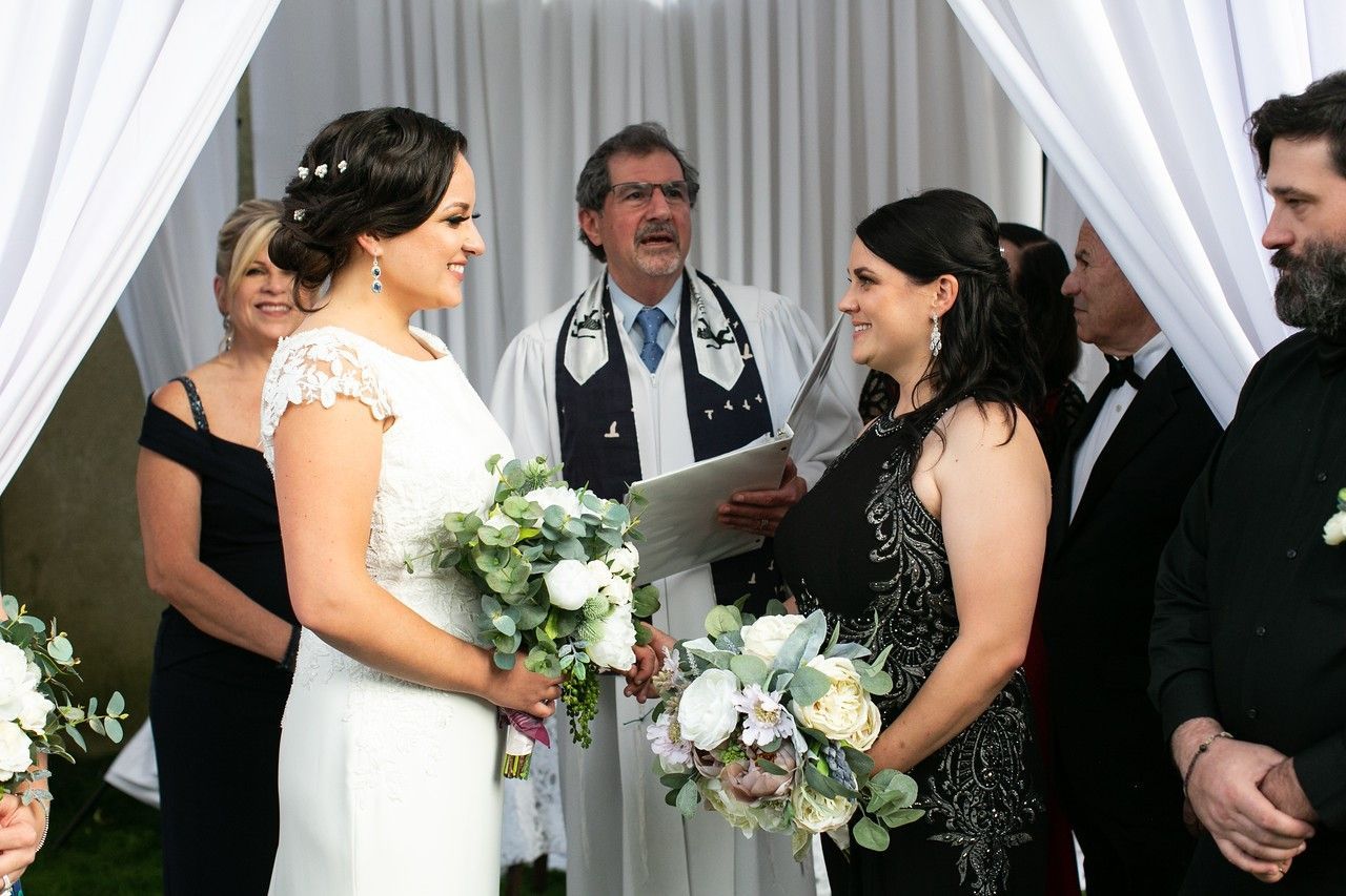 Two women at a wedding ceremony, exchanging vows, in front of an officiant and guests, under white draping.