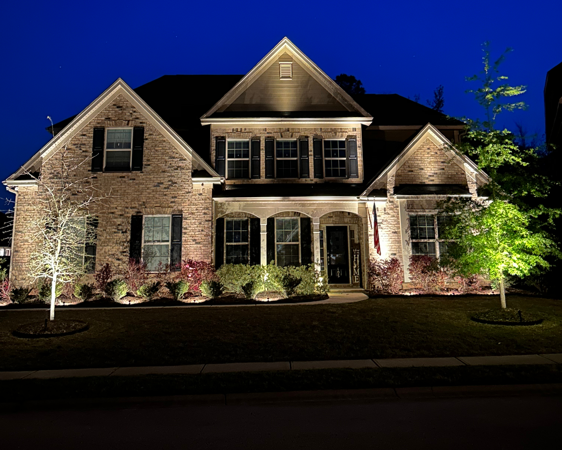 Two-story brick house illuminated at night with landscape lighting; trees and bushes are lit up.