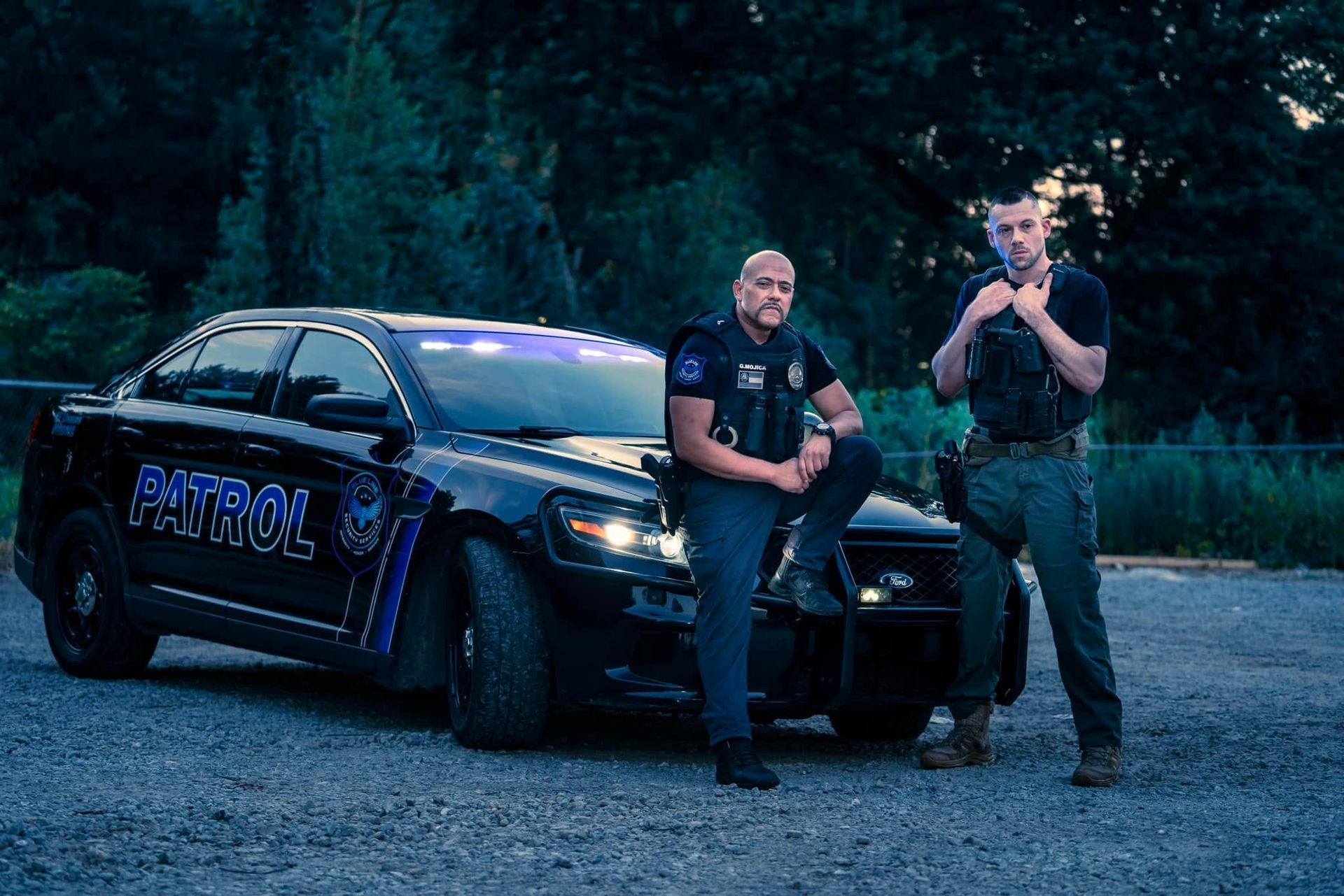 Two police officers stand beside a black police car on a gravel road at dusk.