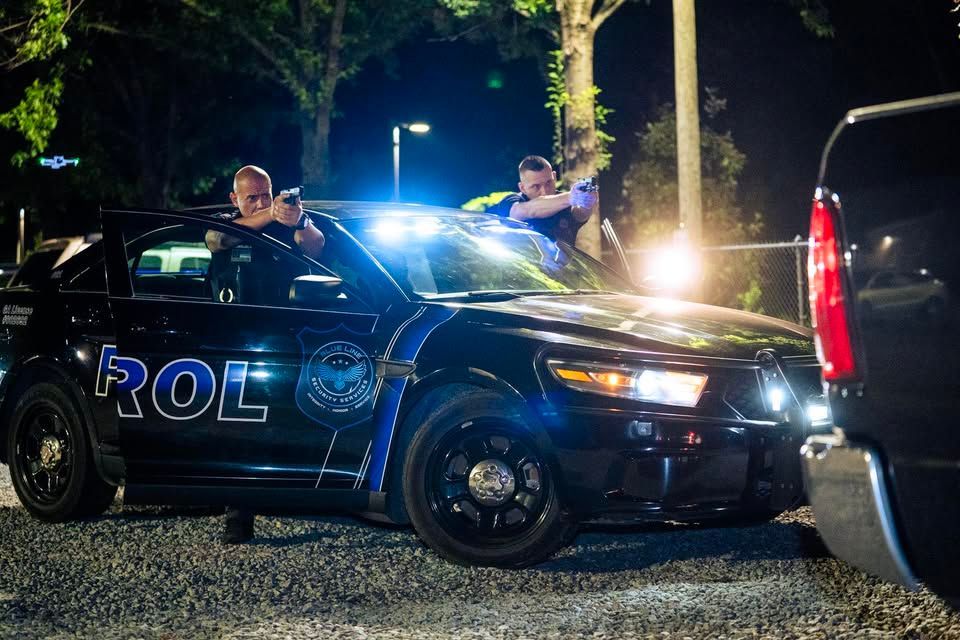 Police officers behind a black patrol car at night, with bright flashing lights and a vehicle in the foreground