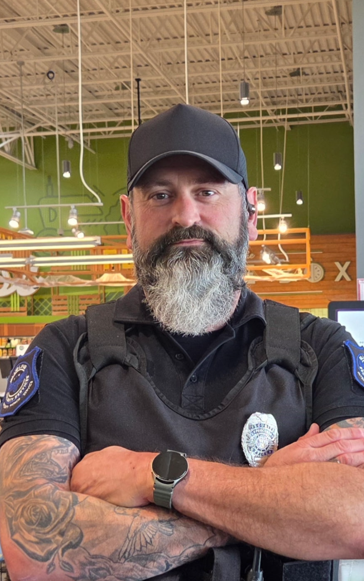 Man in black cap and uniform with tattooed arms, standing arms crossed in a bright indoor setting