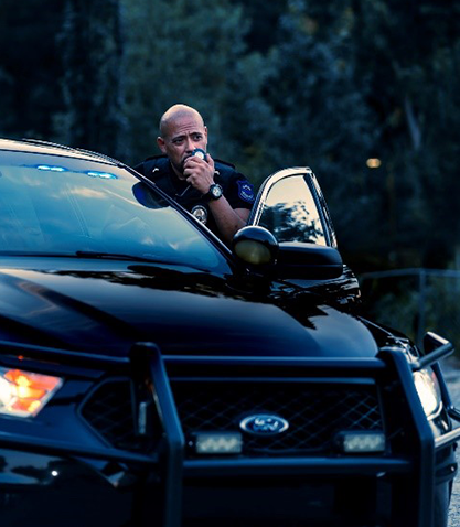 Police officer leaning from a black Ford SUV, speaking into a radio with lights on