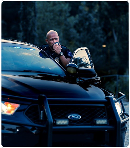 Police officer leaning from a black Ford SUV, speaking into a radio with lights on