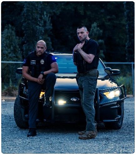 Two police officers stand in front of a black patrol car with headlights on at dusk.