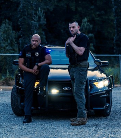 Two police officers stand in front of a black patrol car with headlights on at dusk.