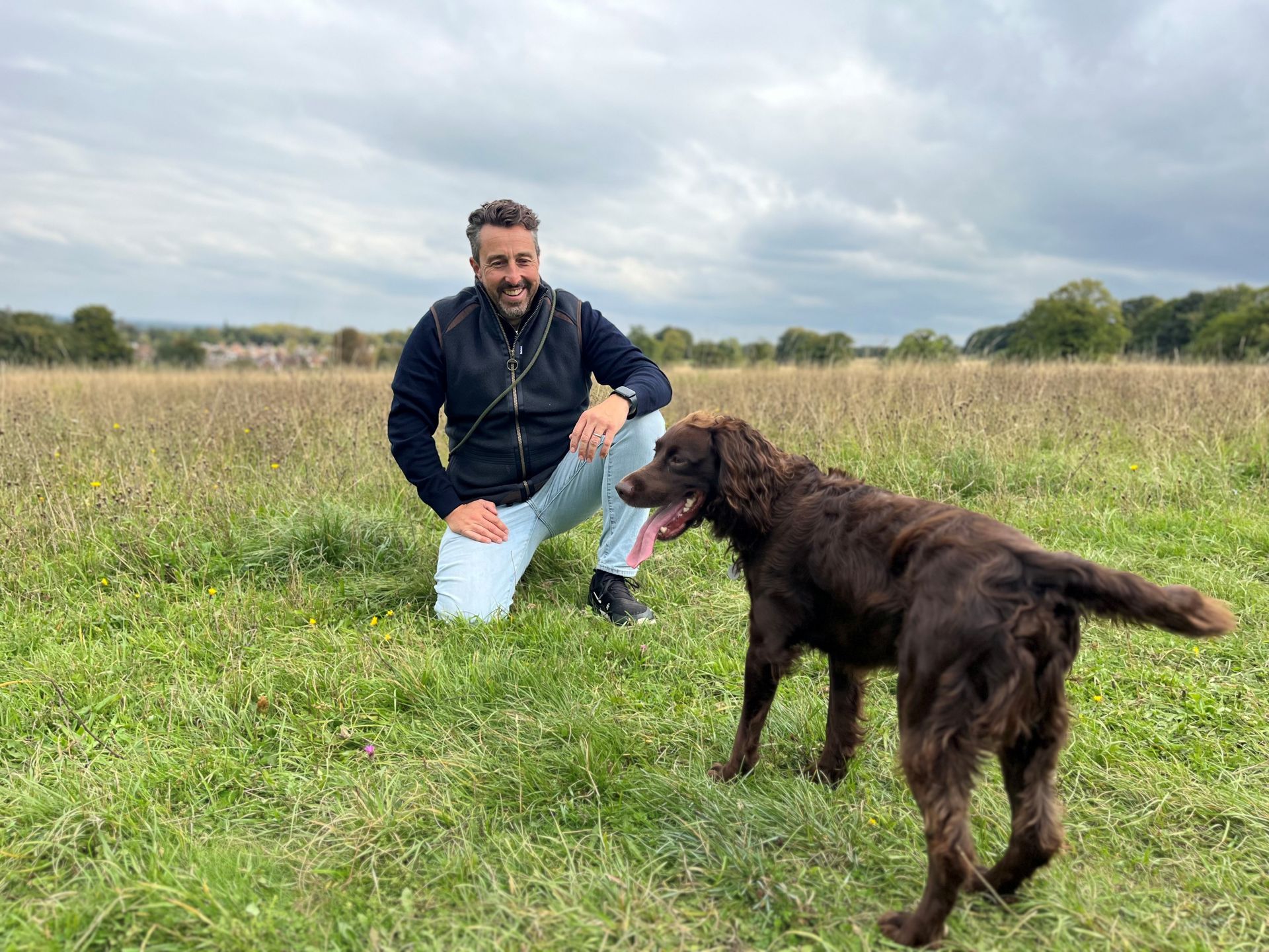 Ecology Recruiter kneeling in field with brown dog, both smiling, overcast sky.
