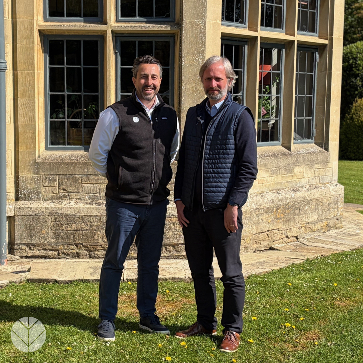 Two men standing outside a stone building, smiling on a sunny day.